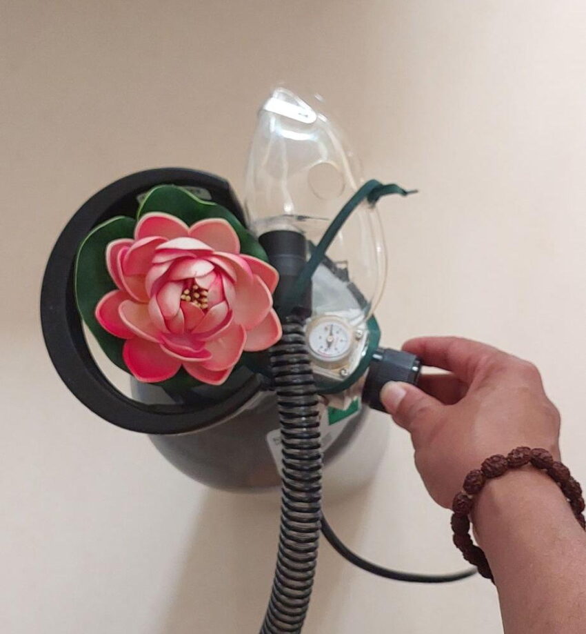 6 litre tank for inhalation with large valve. The tank has a lotos flower placed on top of it. Swamiji's hand with mala bracelet is adjusting the pressure